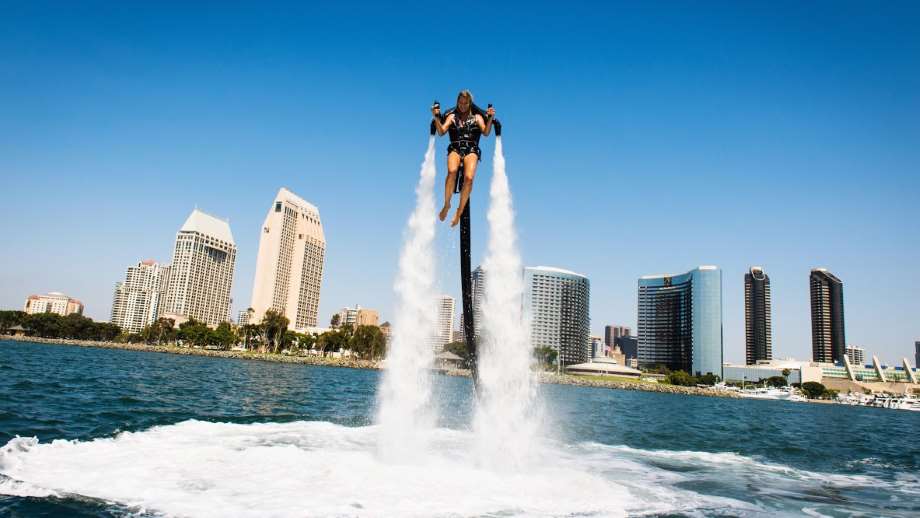 Jetpack by jetpack america shows a girl with a jetpack atop of the water with water shooting out of the pack and a city in the background